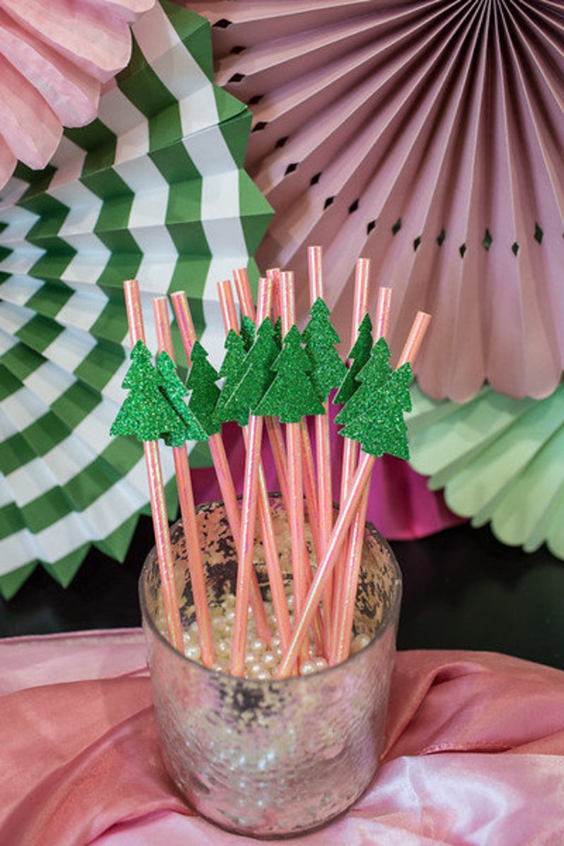 May include: A close-up of a glass jar filled with pink and white striped paper straws with green glitter Christmas tree toppers. The jar is sitting on a pink and white tablecloth.