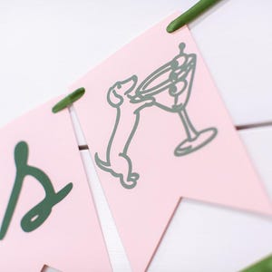 May include: A pink banner with green accents. The banner features a dachshund dog reaching for a martini glass. The letters are in a cursive font. The banner is strung together with green string and is set against a white background.