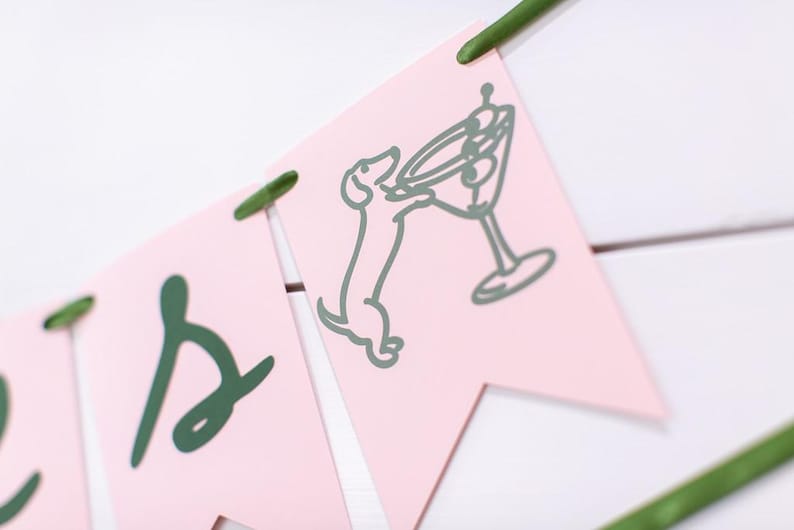 May include: A pink banner with green accents. The banner features a dachshund dog reaching for a martini glass. The letters are in a cursive font. The banner is strung together with green string and is set against a white background.