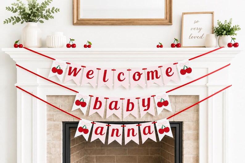 May include: A decorative banner with the words "welcome baby anna" in red letters on pink pennants, adorned with cherry illustrations. The banner is strung across a white mantelpiece, decorated with cherry accents and greenery.