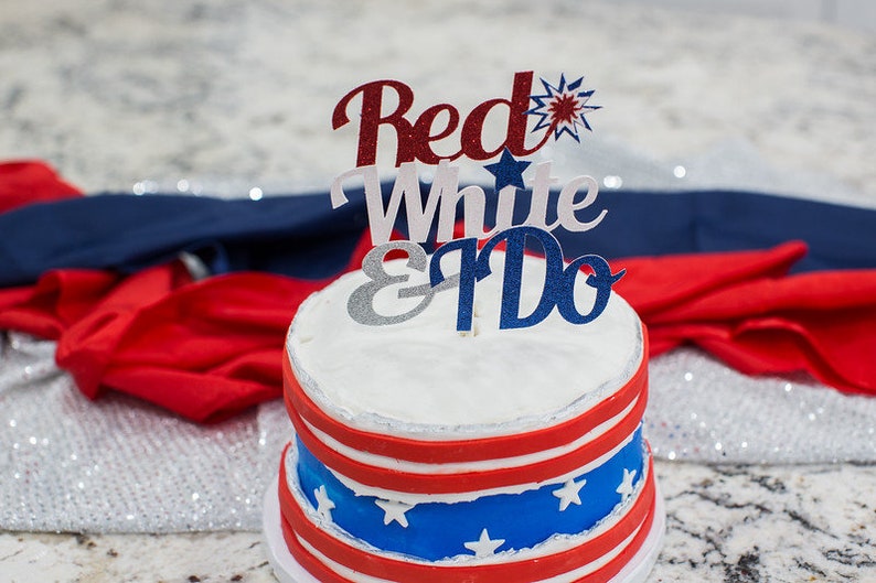 Pode incluir: Um bolo branco de dois andares decorado com listras vermelhas e azuis e estrelas brancas. O bolo &eacute; coberto com um topo de bolo vermelho, branco e azul que diz "Red White & I Do."
