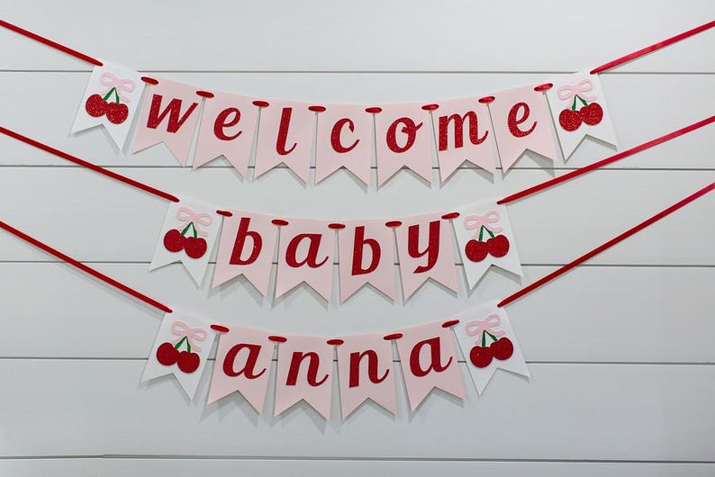 May include: A three-tiered banner with the words "welcome baby anna" in red script on pink pennants. Each pennant is adorned with a red cherry illustration. The banner is strung with red ribbon against a white wood-paneled background.