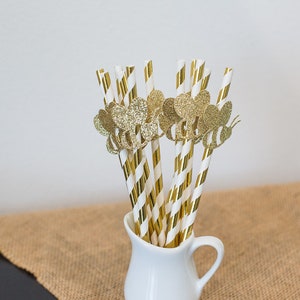 May include: A bunch of gold and white striped paper straws with gold glitter bee toppers in a white pitcher.