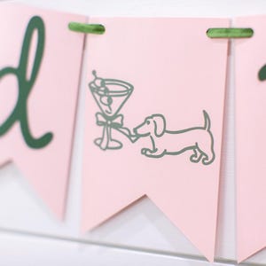 May include: Pink banner with green text and accents. One banner features a martini glass and a dachshund dog illustration. The banner is strung together with green twine. The overall theme is party or celebration.
