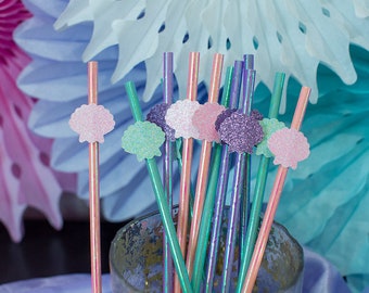 May include: A bunch of colorful straws with glitter seashells attached to the top. The straws are in a clear glass jar. The background is blue and pink.