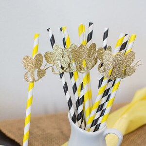 May include: A collection of decorative paper straws in a white pitcher. The straws feature yellow and white stripes, and black and white stripes. Gold glitter bee cutouts adorn the straws, adding a whimsical touch. A yellow cloth is in the background.