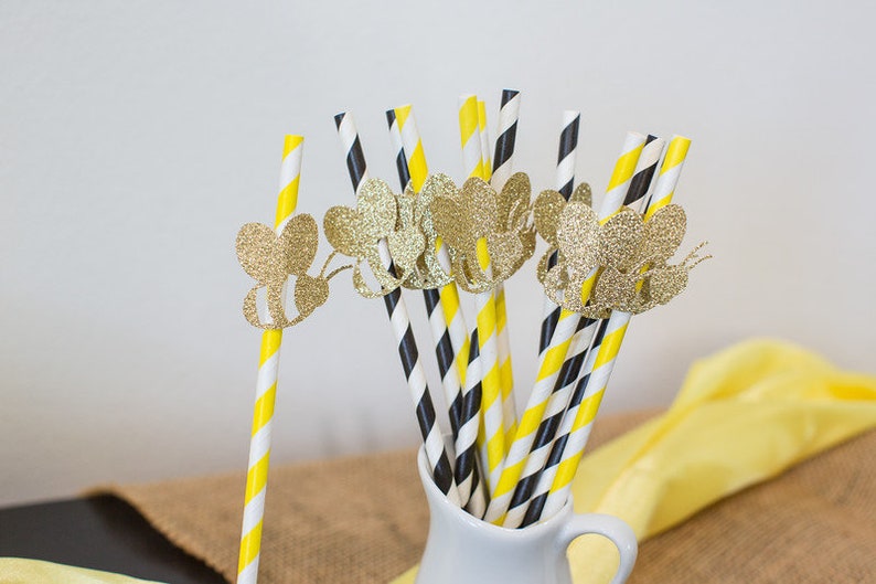 May include: A collection of decorative paper straws in a white pitcher. The straws feature yellow and white stripes, and black and white stripes. Gold glitter bee cutouts adorn the straws, adding a whimsical touch. A yellow cloth is in the background.