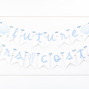 Op de afbeelding: Een decoratieve banner met twee lagen bestaande uit witte wimpels aan een lichtblauw lint. De bovenste rij spelt future met schelpenafbeeldingen en de onderste rij spelt mrs. costa met afbeeldingen van champagneglazen in lichtblauw.