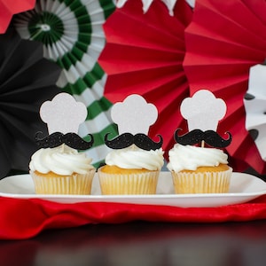 May include: Three white cupcakes with white frosting topped with a black mustache and a white chef's hat. The cupcakes are on a white plate and a red tablecloth. The background is a colorful paper fan backdrop.