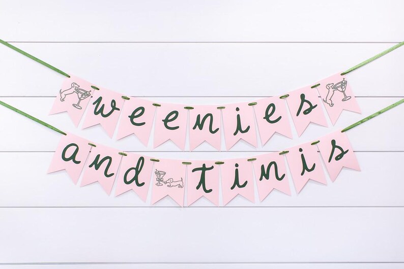May include: Pink banner with the words "weenies and tinis" in dark green script. Each pennant is pink and has a small illustration of a dachshund and a martini glass. The banner is strung on green ribbon.