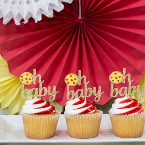 May include: Three yellow and white cupcakes with red frosting and gold glitter "Oh Baby" toppers featuring a yellow and red polka dot design.