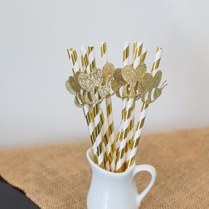 May include: A bunch of gold and white striped paper straws with gold glitter bee toppers in a white pitcher.