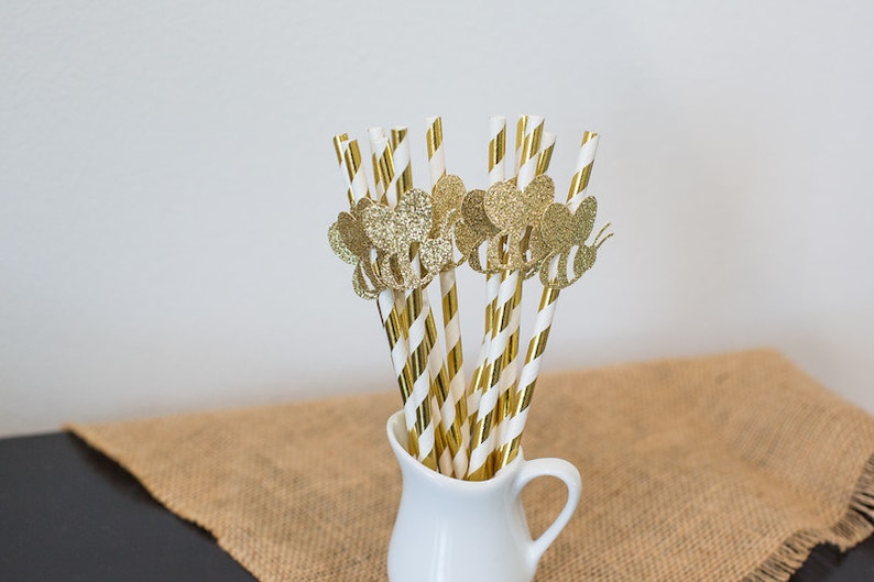 May include: A bunch of gold and white striped paper straws with gold glitter bee toppers in a white pitcher.