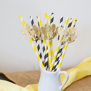 May include: A collection of decorative paper straws in a white pitcher. The straws have yellow and black stripes, and black and white diagonal stripes. Gold glitter butterfly embellishments adorn some of the straws. A yellow cloth and burlap are in the background.