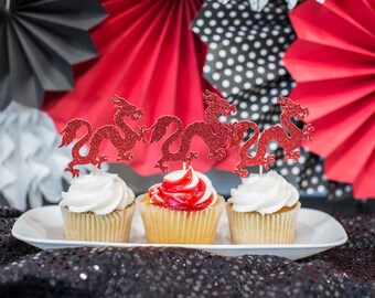May include: Three cupcakes decorated with red glitter dragon toppers. The cupcakes are on a white plate and the background is red, black, and white.