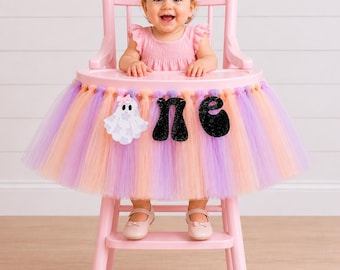 May include: A pink high chair decorated with a multi-coloured tulle skirt in shades of lavender and peach. The skirt features a white ghost cutout with a small pink bow and black glittery letters spelling out the word one on the front.