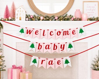 May include: A festive banner with the words "welcome baby rae" in red lettering on pink pennants, adorned with green Christmas trees. The banner is strung across a white fireplace, decorated with Christmas trees and pink ornaments.