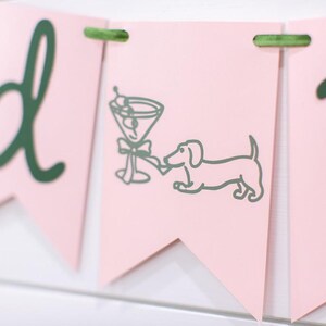 May include: Pink banner with green lettering and accents. One banner features a martini glass and a dachshund dog illustration. The banner is strung together with green twine.