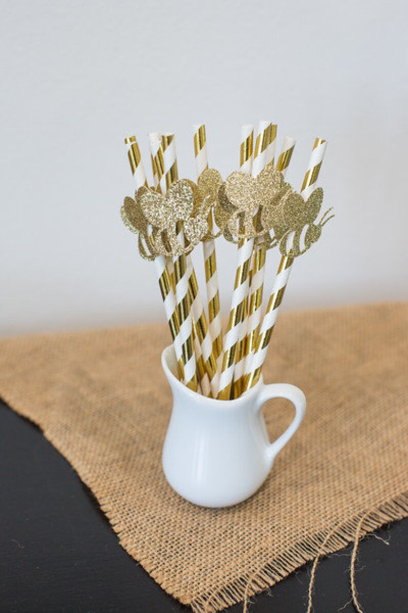 May include: Gold and white striped paper straws with glitter bee toppers in a white pitcher. The pitcher is sitting on a brown burlap surface.