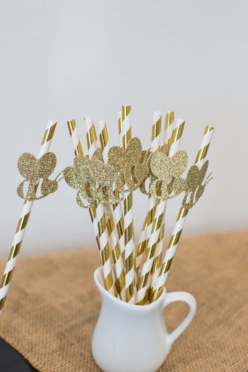 May include: Gold glitter straws with a heart-shaped design. The straws are white and gold striped and are in a white pitcher.