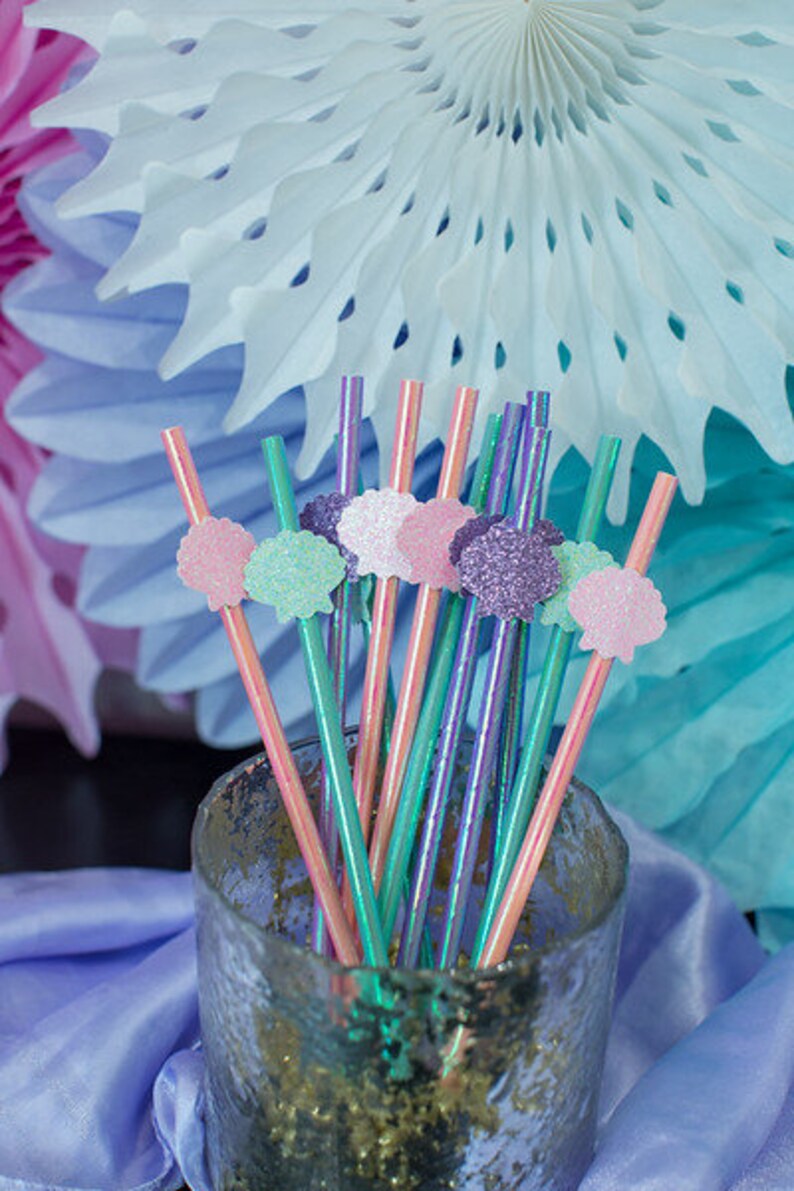 May include: A glass jar filled with colorful iridescent straws with decorative seashell toppers in pink, green, and purple. The straws are perfect for a mermaid-themed party.