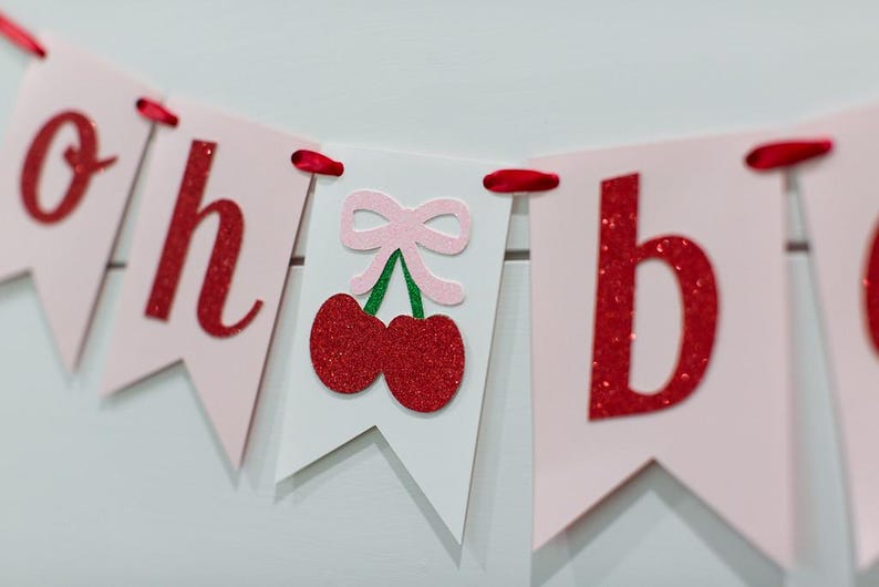 May include: A decorative banner featuring light pink and white pennants strung together with red ribbon. The pennants display red glittery letters spelling out "oh" and "b", with a central white pennant showing a pair of red glittery cherries.