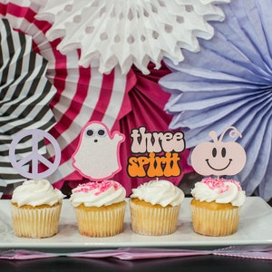 May include: Four cupcakes decorated with pink frosting and various toppers. The toppers include a peace sign, a ghost, the words "three spirit", and a smiling pumpkin. The cupcakes are on a white plate in front of a background of pink, white, and purple paper fans.