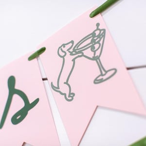 May include: Pink banner with green lettering and illustrations. One banner features a dachshund dog reaching for a martini glass. The banner is strung together with green string and is set against a white background.