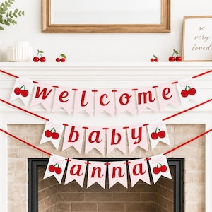 May include: A decorative banner with the words "welcome baby anna" in red letters on pink pennants, adorned with cherry illustrations. The banner is strung across a white mantelpiece, decorated with cherry accents and greenery.