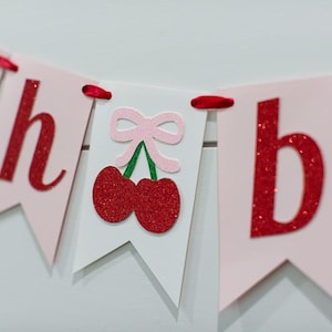 May include: A decorative banner featuring light pink and white pennants strung together with red ribbon. The pennants display red glittery letters spelling out "oh" and "b", with a central white pennant showing a pair of red glittery cherries.