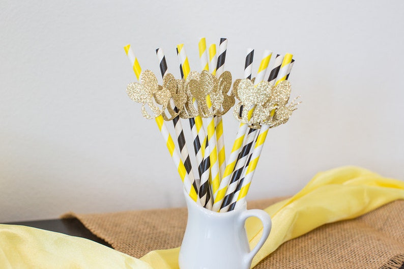 May include: A collection of decorative paper straws in a white pitcher. The straws have yellow and black stripes, and black and white diagonal stripes. Gold glitter butterfly embellishments adorn some of the straws. A yellow cloth and burlap are in the background.