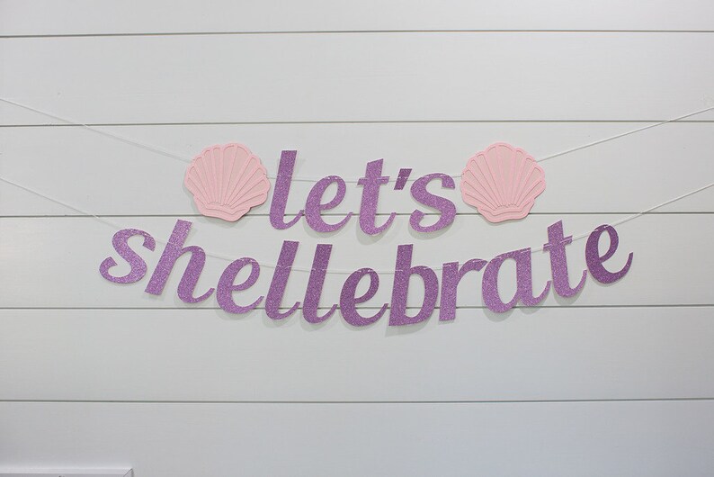 May include: Purple glitter banner with the text "Let's Shellebrate" and two pink seashells on a white wood background.