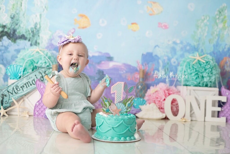 May include: A baby girl wearing a light blue dress and a purple flower crown is sitting in front of a cake decorated with a mermaid tail and a number one. The cake is on a white plate and there is a wooden spoon in the baby's hand. The background is a blue and green ocean scene with a sign that says "Mermaids".