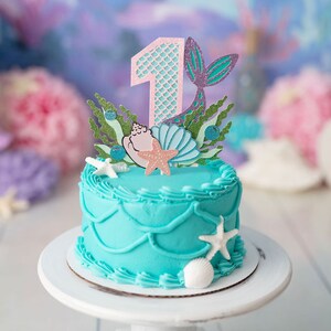 May include: A turquoise frosted cake with a mermaid tail and seashell topper. The cake is decorated with white frosting and has a number one on top. The cake is on a white cake stand.