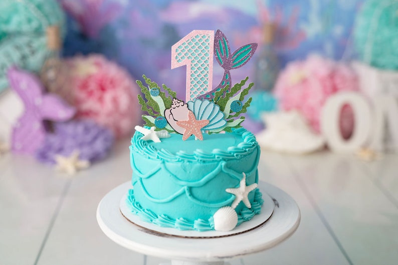 May include: A turquoise frosted cake with a mermaid tail and seashell topper. The cake is decorated with white frosting and has a number one on top. The cake is on a white cake stand.