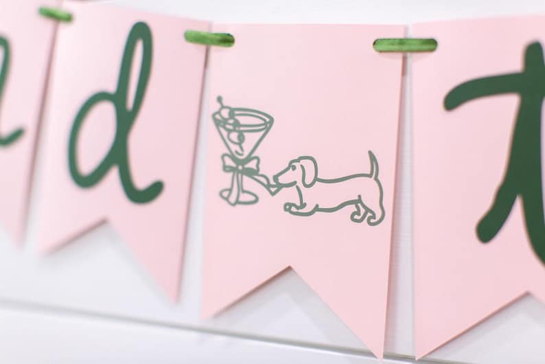 May include: Pink banner with green text and accents. One banner features a martini glass and a dachshund dog illustration. The banner is strung together with green twine. The overall theme is party or celebration.