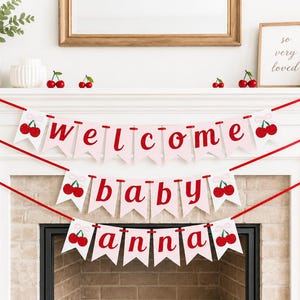 May include: A decorative banner with the words "welcome baby anna" in red letters on pink pennants, adorned with cherry illustrations. The banner is strung across a white mantelpiece, decorated with cherry accents and greenery.