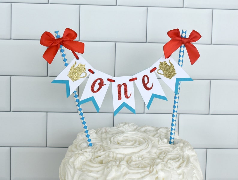 May include: A white cake with white frosting decorated with a red, white, and blue banner that says "one" with a gold teapot on each end. The banner is attached to two blue and white striped straws with red bows on top.