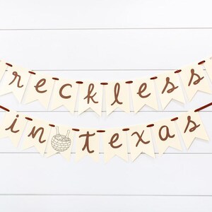 May include: A banner with the words "reckless in texas" in brown script on cream-colored pennants. The pennants are strung on brown ribbons and hang against a white, horizontal-plank background. The banner includes cocktail and basket illustrations.