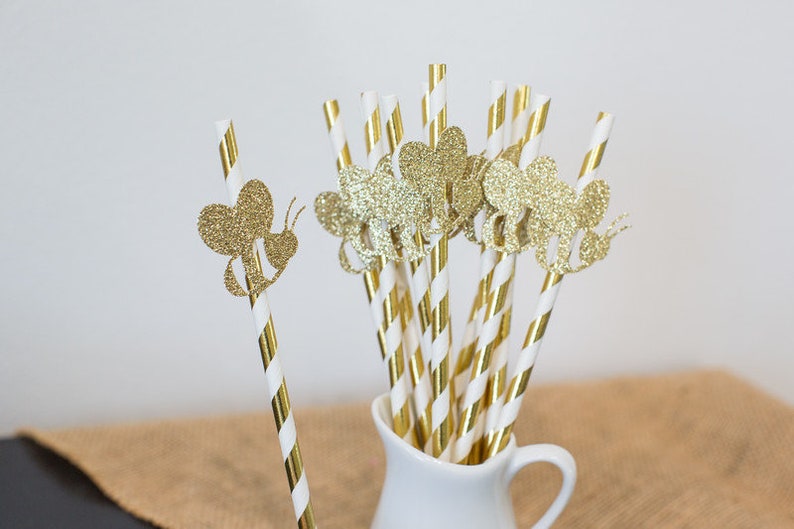 May include: Gold and white striped paper straws with gold glitter bee cutouts. The straws are in a white pitcher.