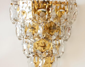 May include: A decorative wall sconce with a tiered design. The sconce features clear and gold-colored crystal elements. The crystals are arranged in a layered pattern, creating a sparkling and elegant appearance. The sconce is mounted on a white wall.