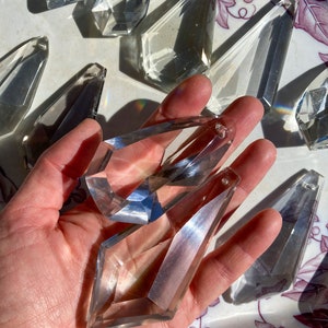 Puede incluir: Una mano sostiene dos prismas de cristal transparentes, cada uno con una forma facetada, similar a un diamante. Los prismas están sobre una superficie blanca con un patrón floral.
