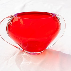 May include: A red glass vase with two clear glass handles. The vase is round and has a wide opening.