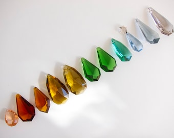 May include: A rainbow assortment of crystal teardrop pendants in various colours, including amber, green, and blue. The teardrop pendants are arranged in a line on a white background.