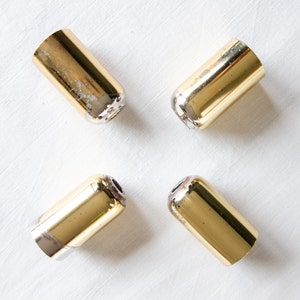 Könnte beinhalten: Vier zylindrische, goldfarbene Metallobjekte mit silberfarbenen Basen sind diagonal auf einer weißen Oberfläche angeordnet. Die Objekte weisen ein leicht abgenutztes Aussehen mit Verfärbungen auf der Goldoberfläche auf.