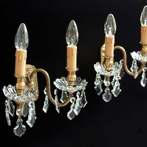 May include: Four vintage wall sconces with clear crystal detailing and candle-shaped bulbs. The sconces have a gold finish and are set against a black backdrop. Each sconce features a decorative crystal base and hanging crystals.