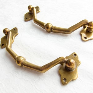 Könnte beinhalten: Zwei antike Messing-Türgriffe mit dekorativem Design. Die Griffe haben eine abgerundete Form und sind an einer quadratischen Platte befestigt.