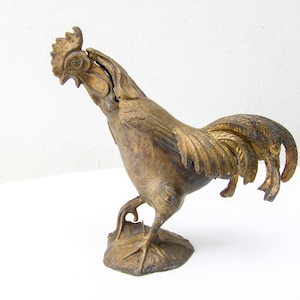 Puede incluir: Una figurita de gallo de metal de color dorado con un diseño detallado. El gallo está de pie con la cabeza girada hacia un lado y las plumas de la cola extendidas.