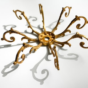 Op de afbeelding: Een gouden metalen kroonluchterarmatuur met zes sierlijke armen, die elk eindigen in een decoratieve krul. De armatuur heeft een centrale ronde basis met een gat in het midden.