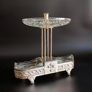 May include: A two-tiered serving stand featuring a rectangular silver-toned metal base with floral relief detailing and two glass inserts, supported by four vertical metal rods holding a circular, scalloped-edge clear glass top dish.